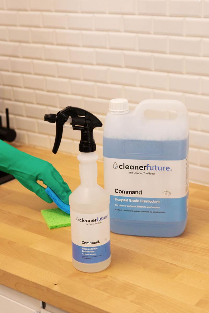 A Cleaner Future Command 5 litre container next to a labelled Cleaner Future dispenser spray bottle on a bench, with a gloved hand and cloth